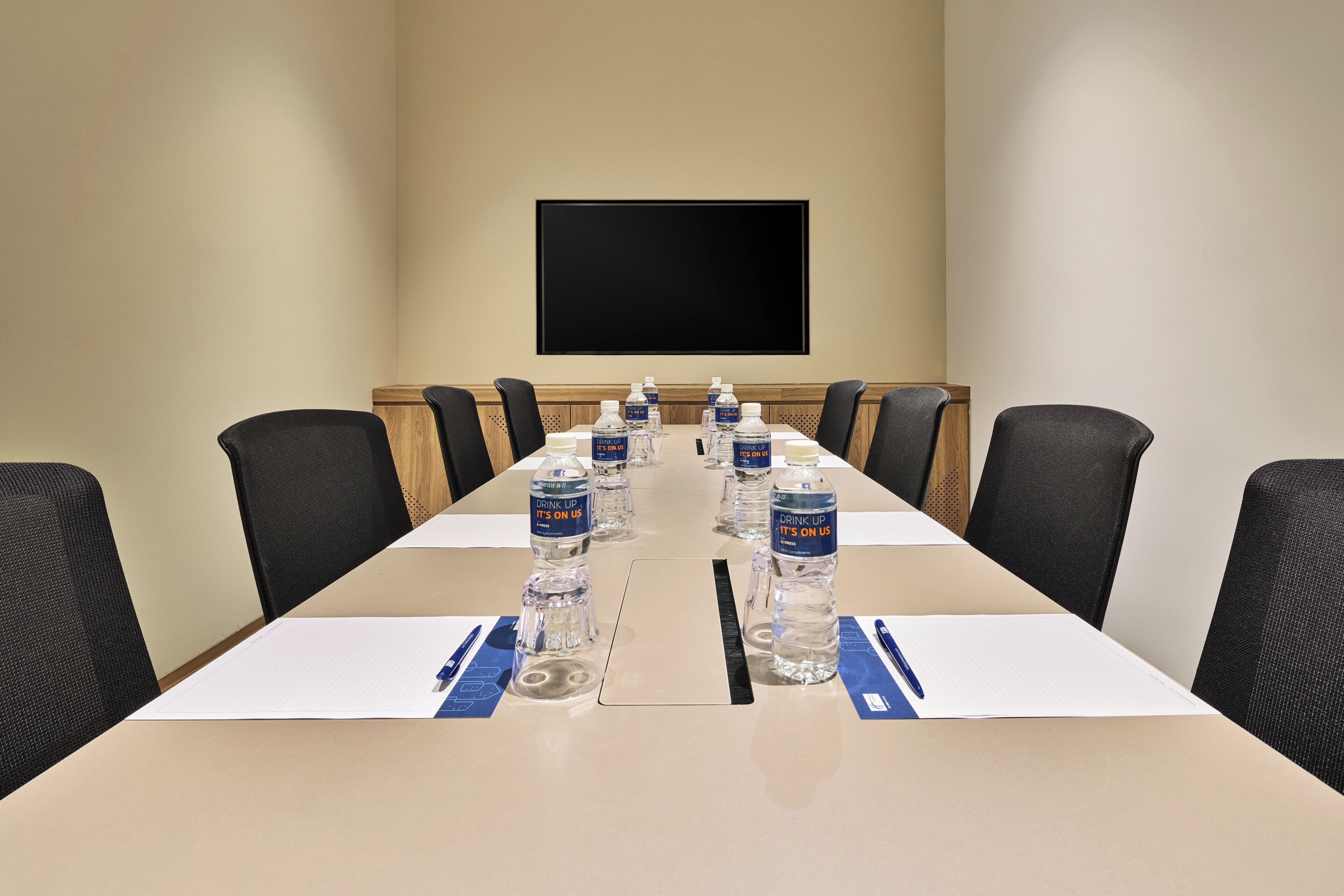 A conference room with a long table, black chairs, and water bottles labeled 'DRINK UP IT'S ON US'.