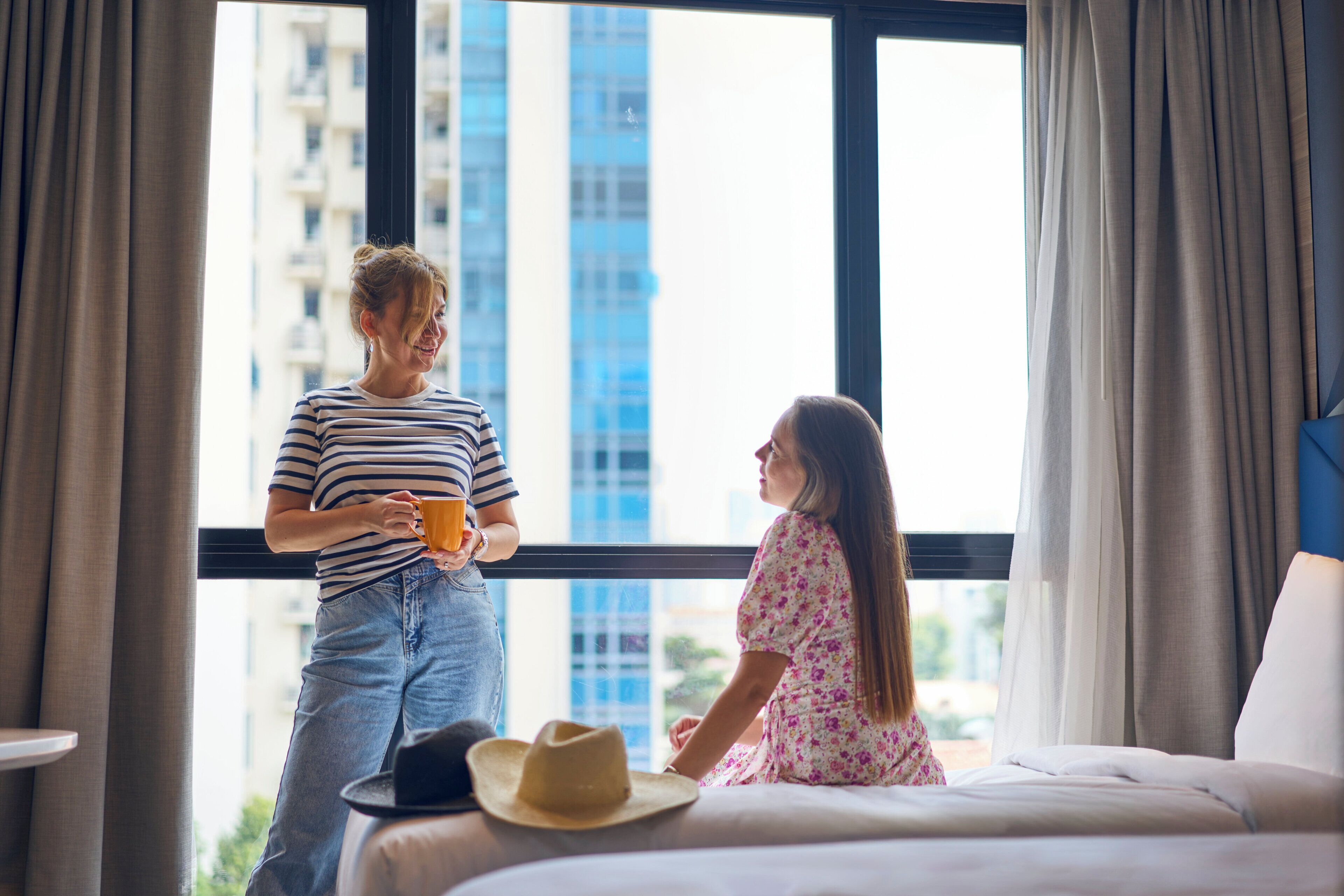 A hotel room with a large window, a bed, and a hat on the bed.