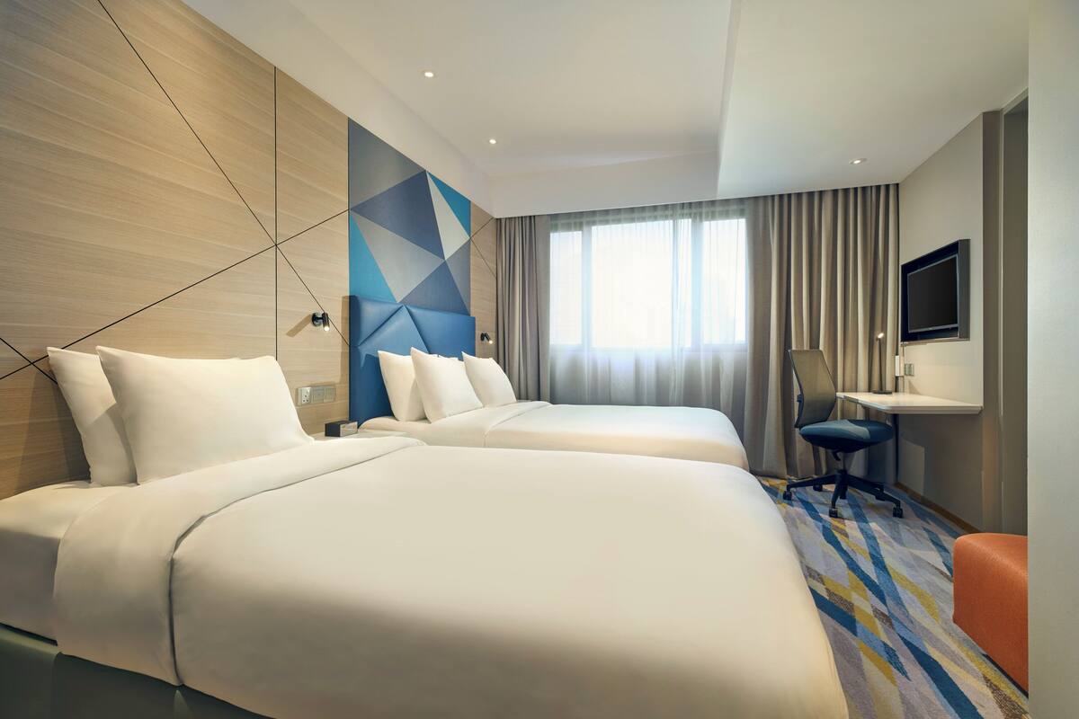 Une chambre d’hôtel avec deux lits, un bureau, une chaise, une télévision et une grande fenêtre avec des rideaux.