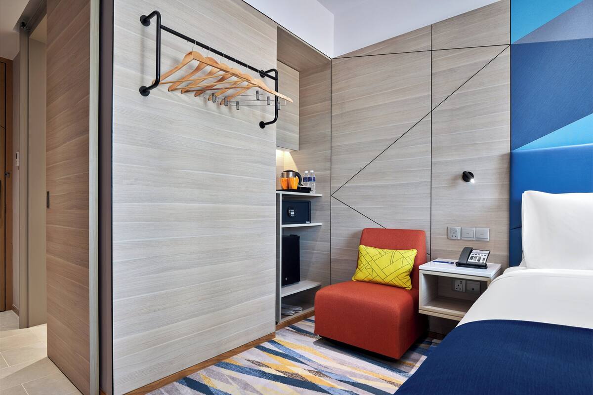 Une chambre d’hôtel avec un lit, une chaise rouge, une petite table avec un téléphone, une étagère avec des articles de toilette et un porte-serviettes fixé au mur.
