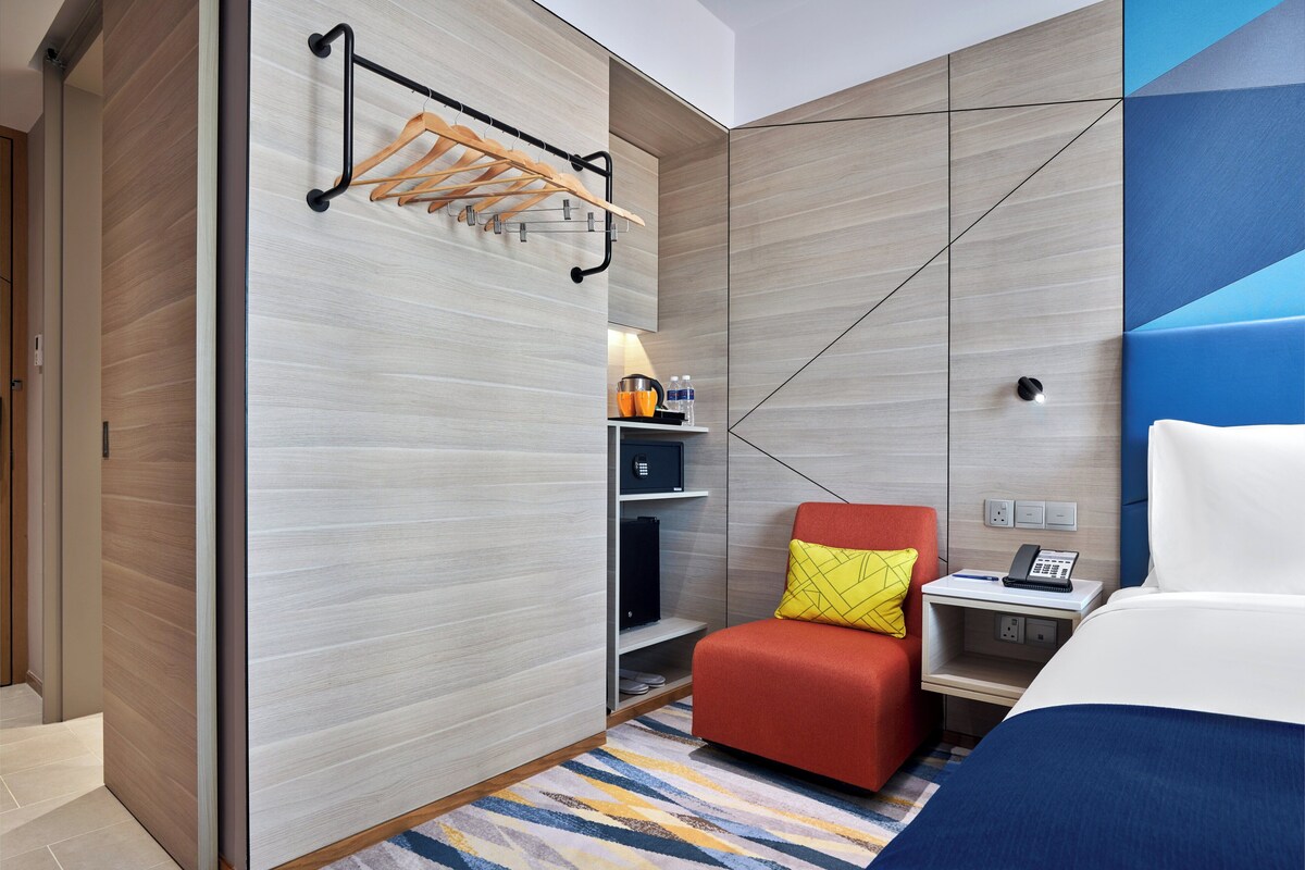 Une chambre d’hôtel avec un lit, une chaise rouge, une petite table avec un téléphone, une étagère avec des articles de toilette et un porte-serviettes fixé au mur.