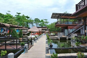Un resort con un paseo de madera que lleva a una piscina, rodeado de abundante vegetación y construcciones con techo de paja.