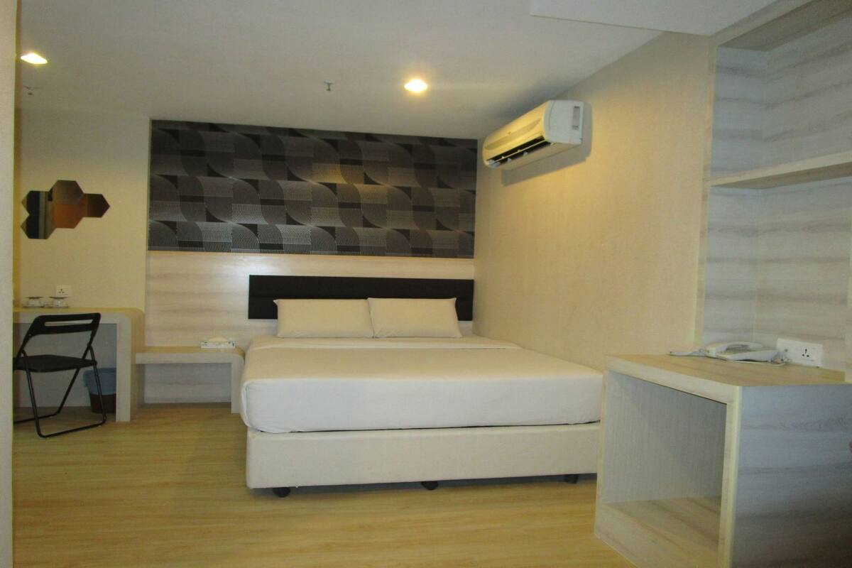 A compact hotel room with a bed, a desk, a chair, and an air conditioning unit.