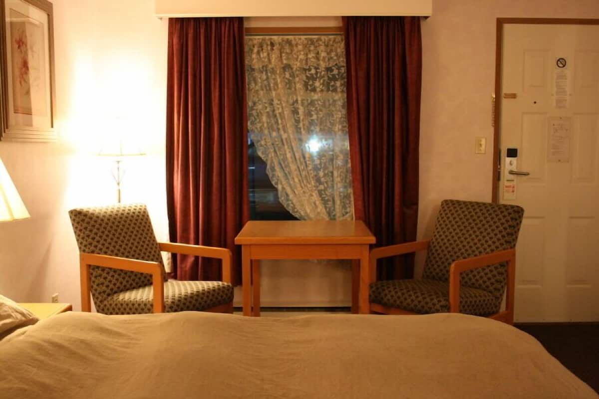 A hotel room with a bed, two armchairs, a small wooden table, and a window with curtains.