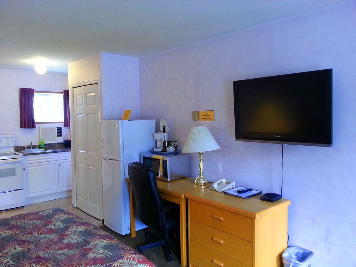 Una habitación de hotel compacta con kitchenette, un escritorio con lámpara, televisor de pantalla plana y cama con colcha estampada.