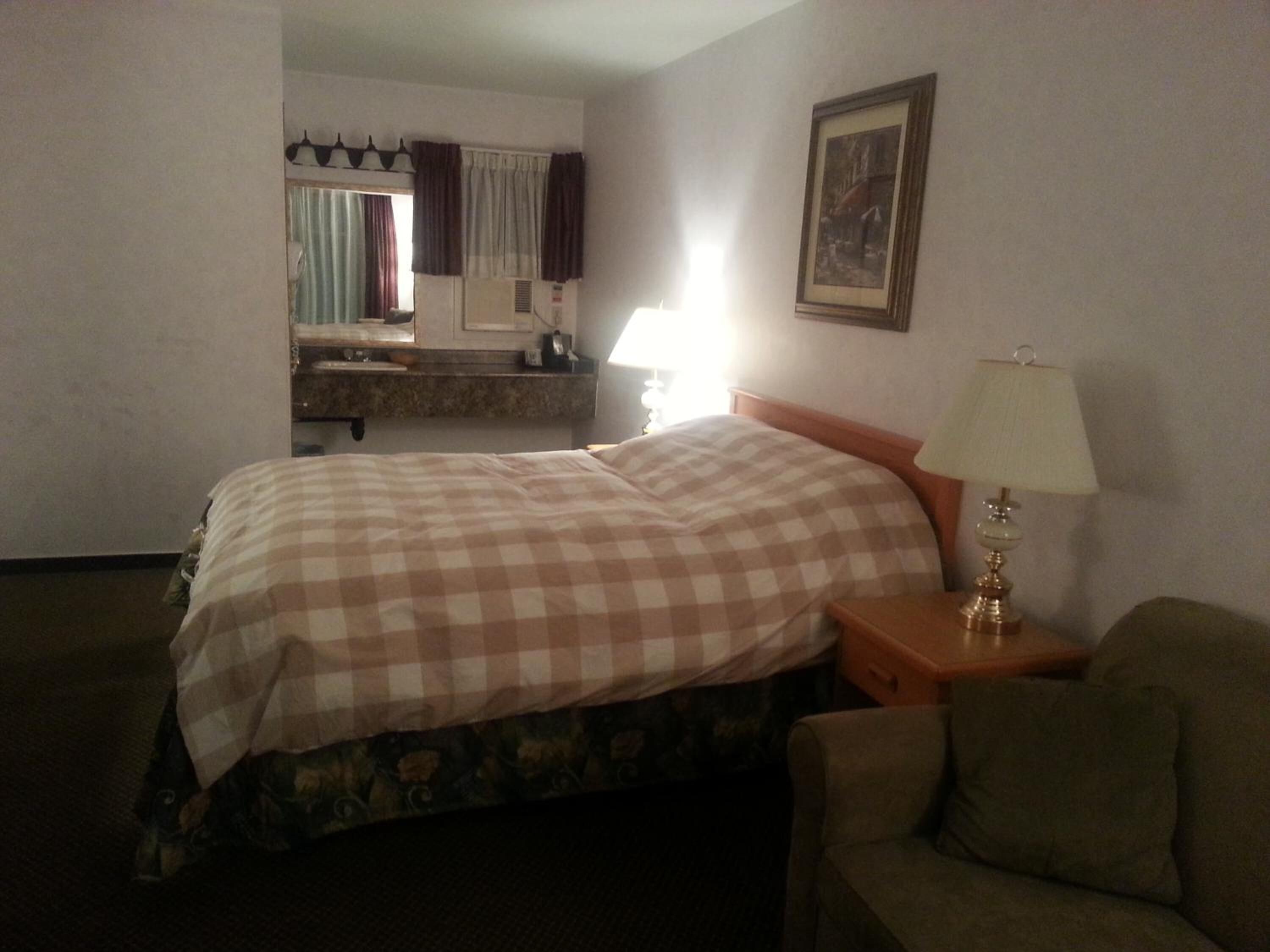 A hotel room with a bed, a nightstand with a lamp, a chair, a bathroom with a mirror, and a framed picture on the wall.