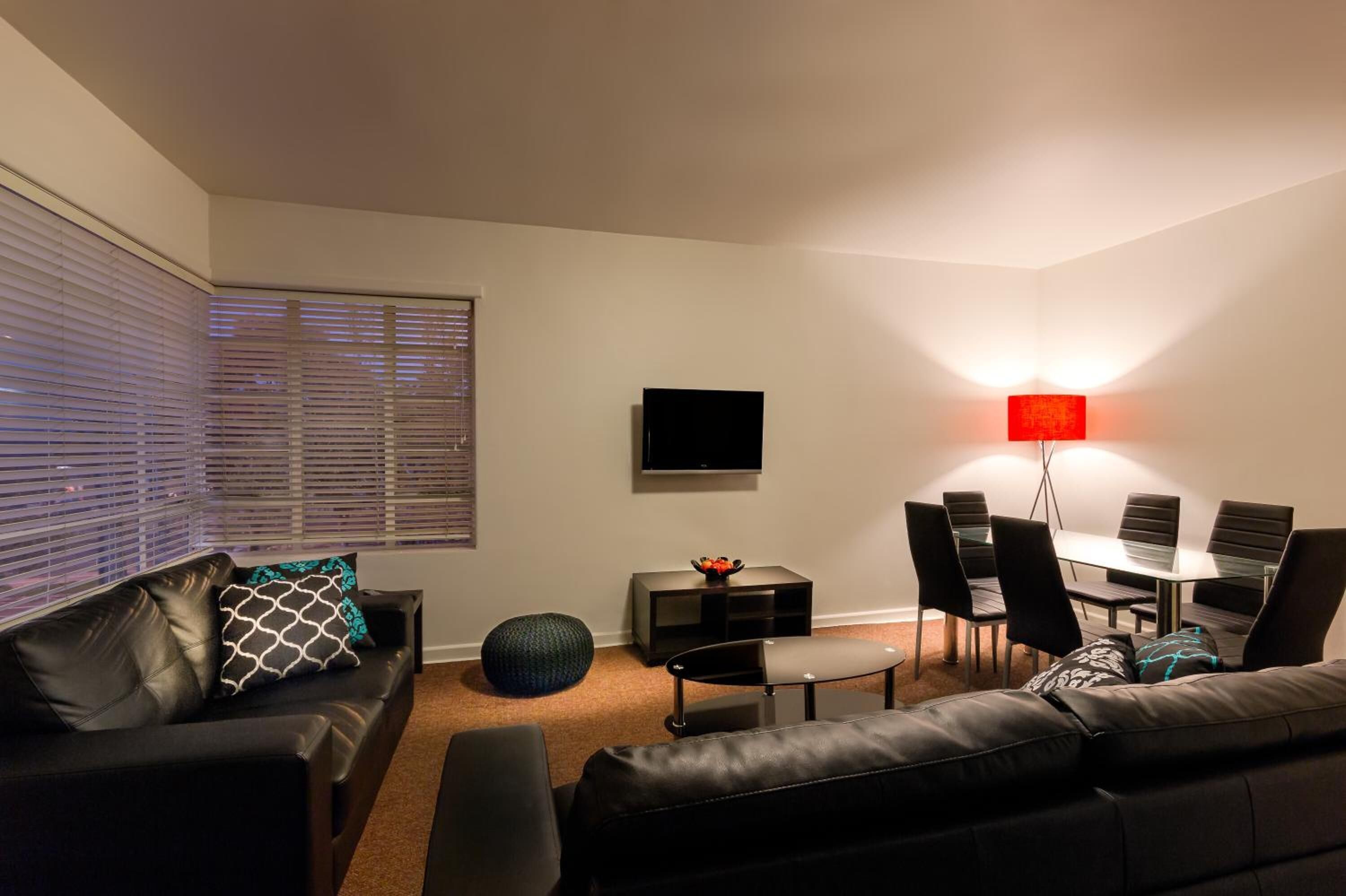 A modern living room with a black leather sofa, a glass dining table with chairs, a flat-screen TV mounted on the wall, and a red floor lamp.