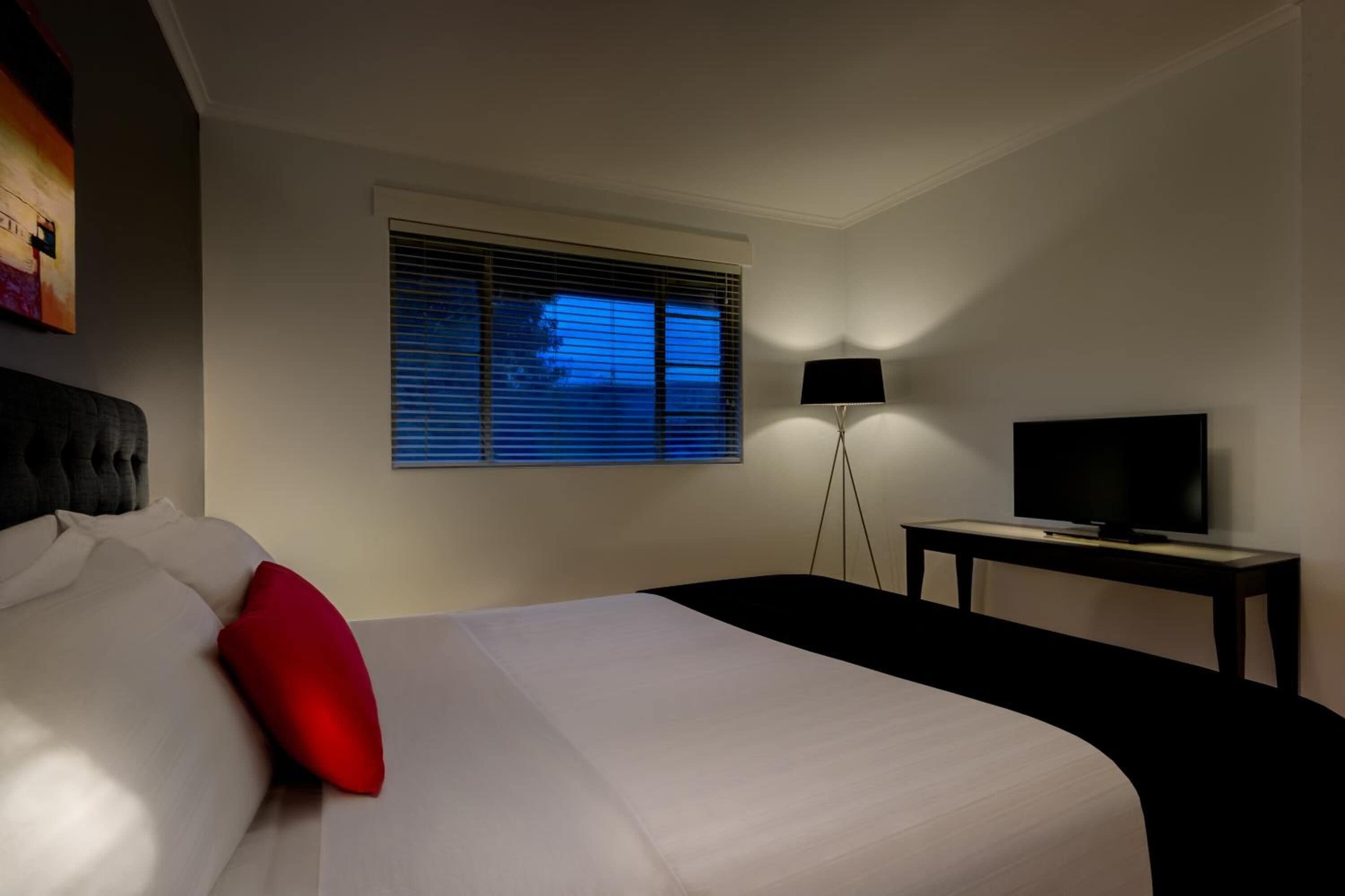 Une chambre d’hôtel avec un lit, un téléviseur posé sur un meuble, une lampe sur pied et une fenêtre avec des stores.