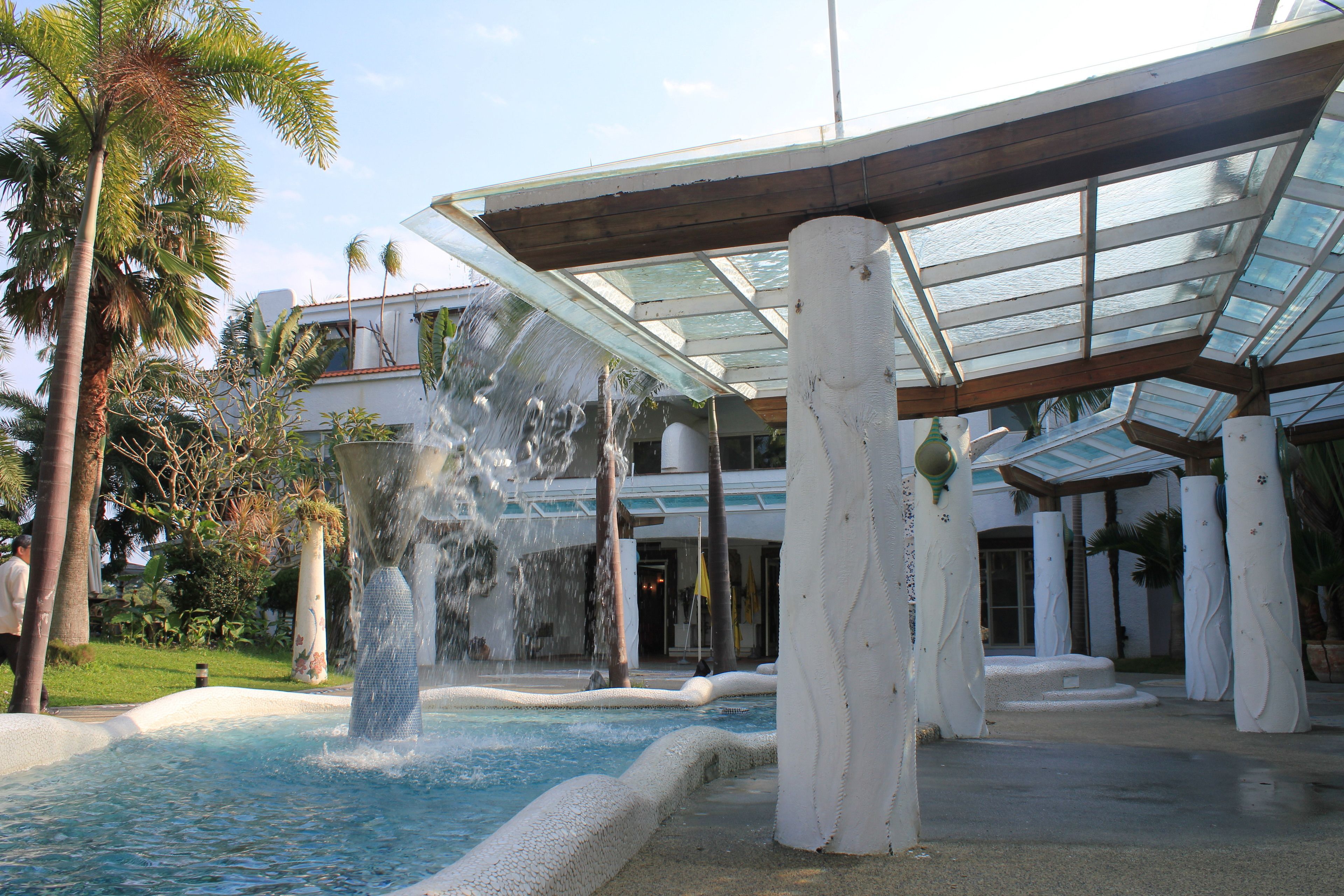 A pool with a waterfall feature, surrounded by palm trees and a covered area with columns.