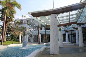 Una piscina con caída de agua, rodeada de palmeras y una zona cubierta con columnas.