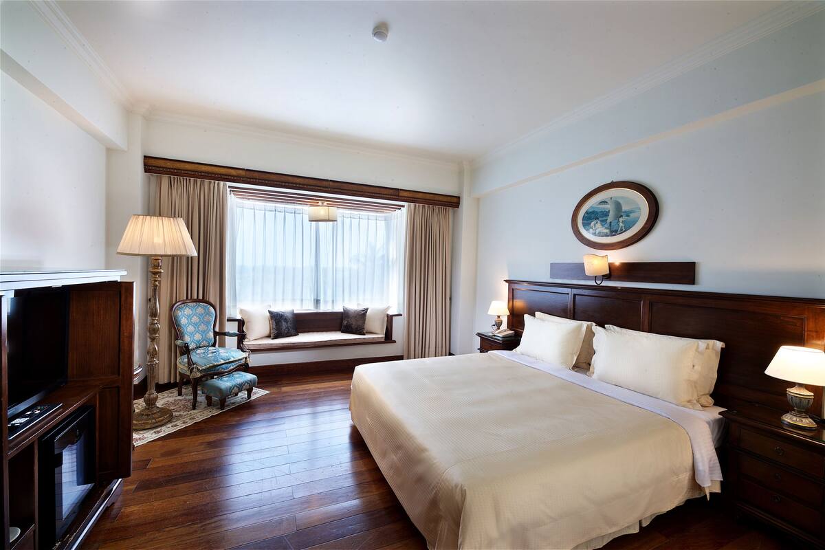 Ein Hotelzimmer mit einem großen Bett, einer Sitzecke mit Sessel und Kissen, einem Fernseher und einem Fenster mit Vorhängen.