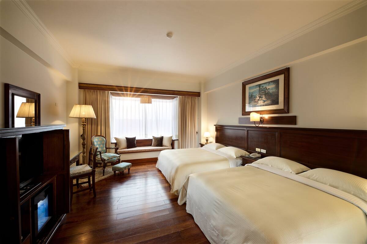 Ein Hotelzimmer mit zwei Betten, einem Fernseher, einem Sessel, einem Fenster mit Vorhängen und einem Bild an der Wand.