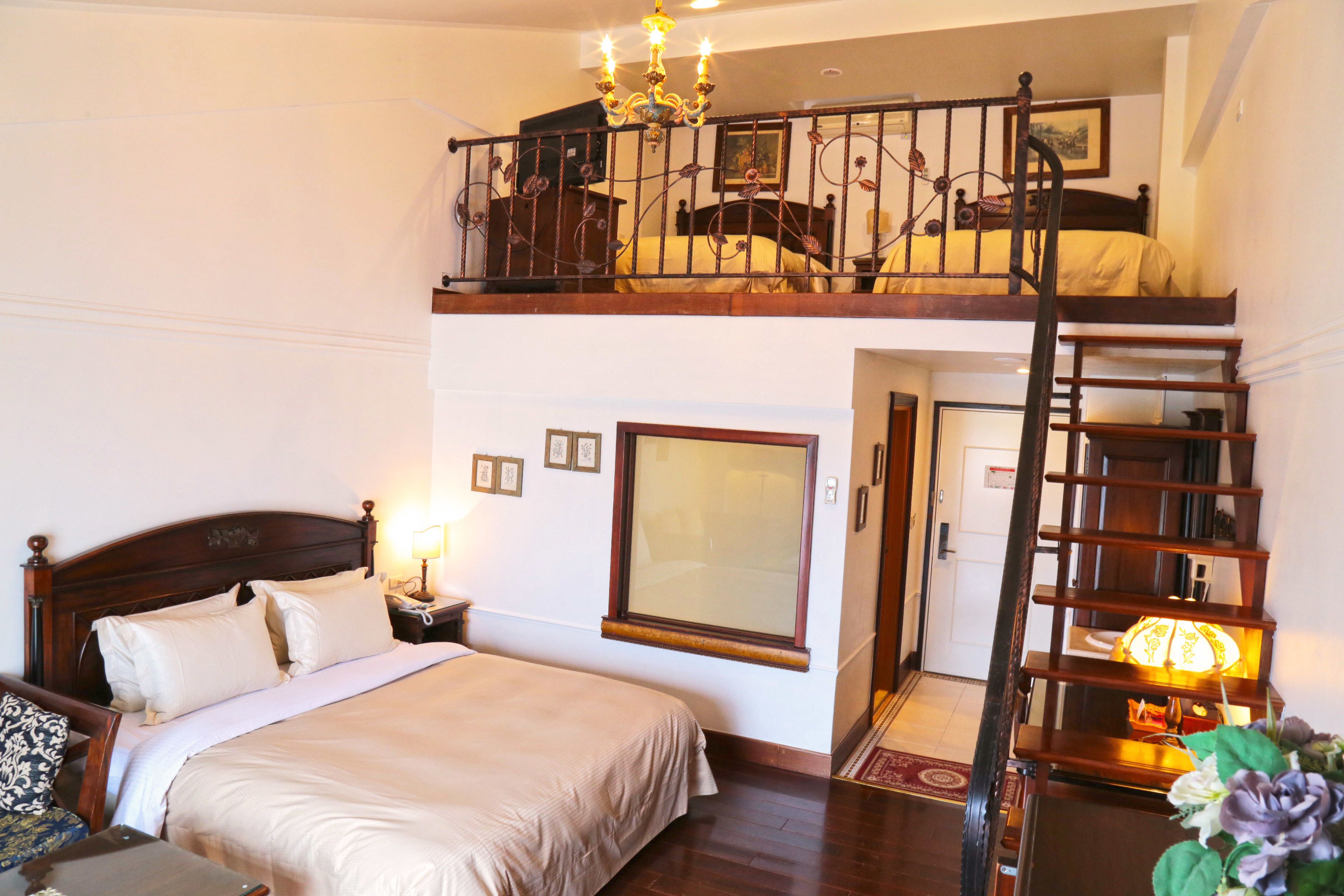 A bedroom with a large bed, a nightstand, a dresser, a chandelier, a balcony with a railing, and a staircase.