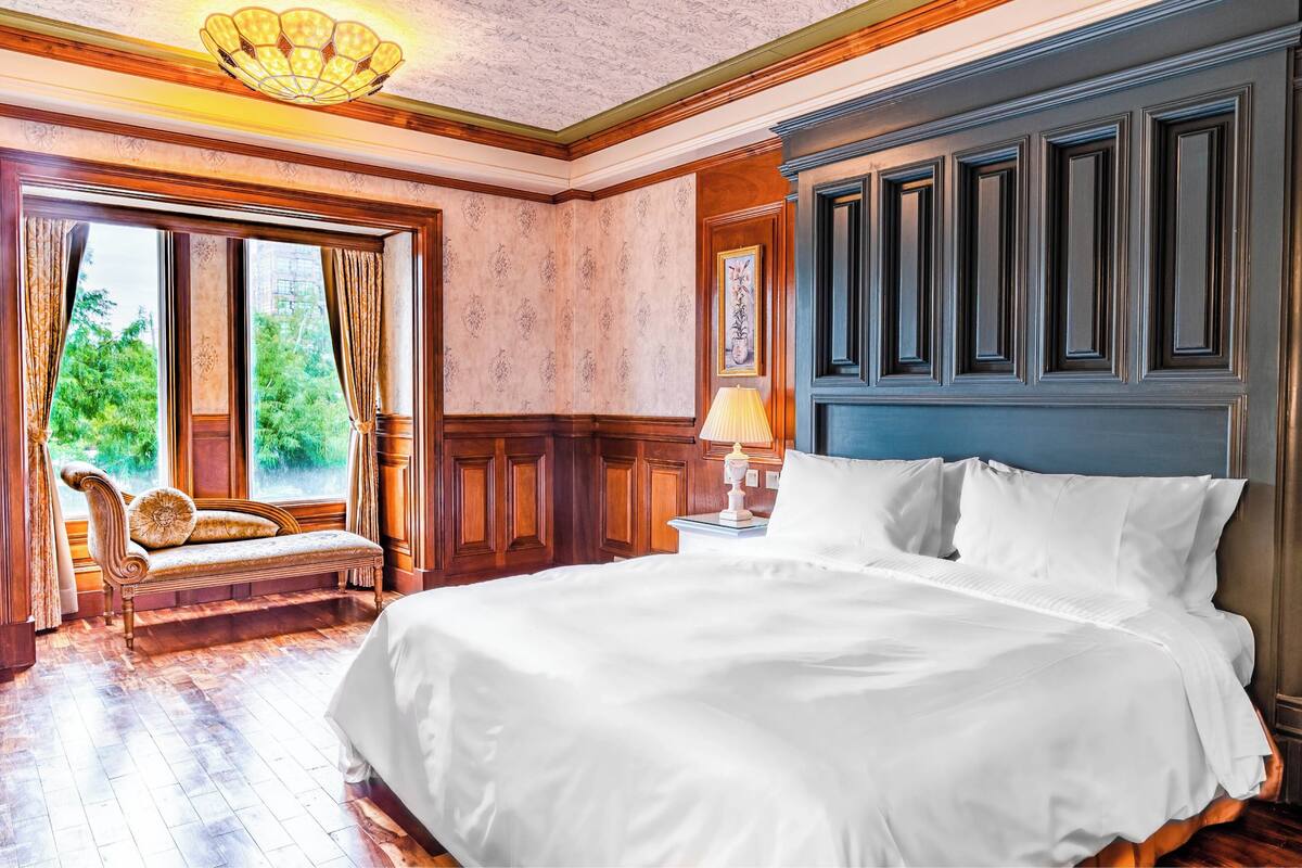 Ein großes Bett mit weißen Leinen, eine Holzbank, ein Nachttisch mit Lampe und ein großes Fenster mit Vorhängen.