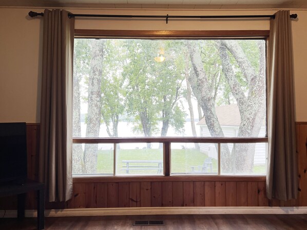 A room with a large window, wooden paneled walls, and a view of trees and a grassy area.