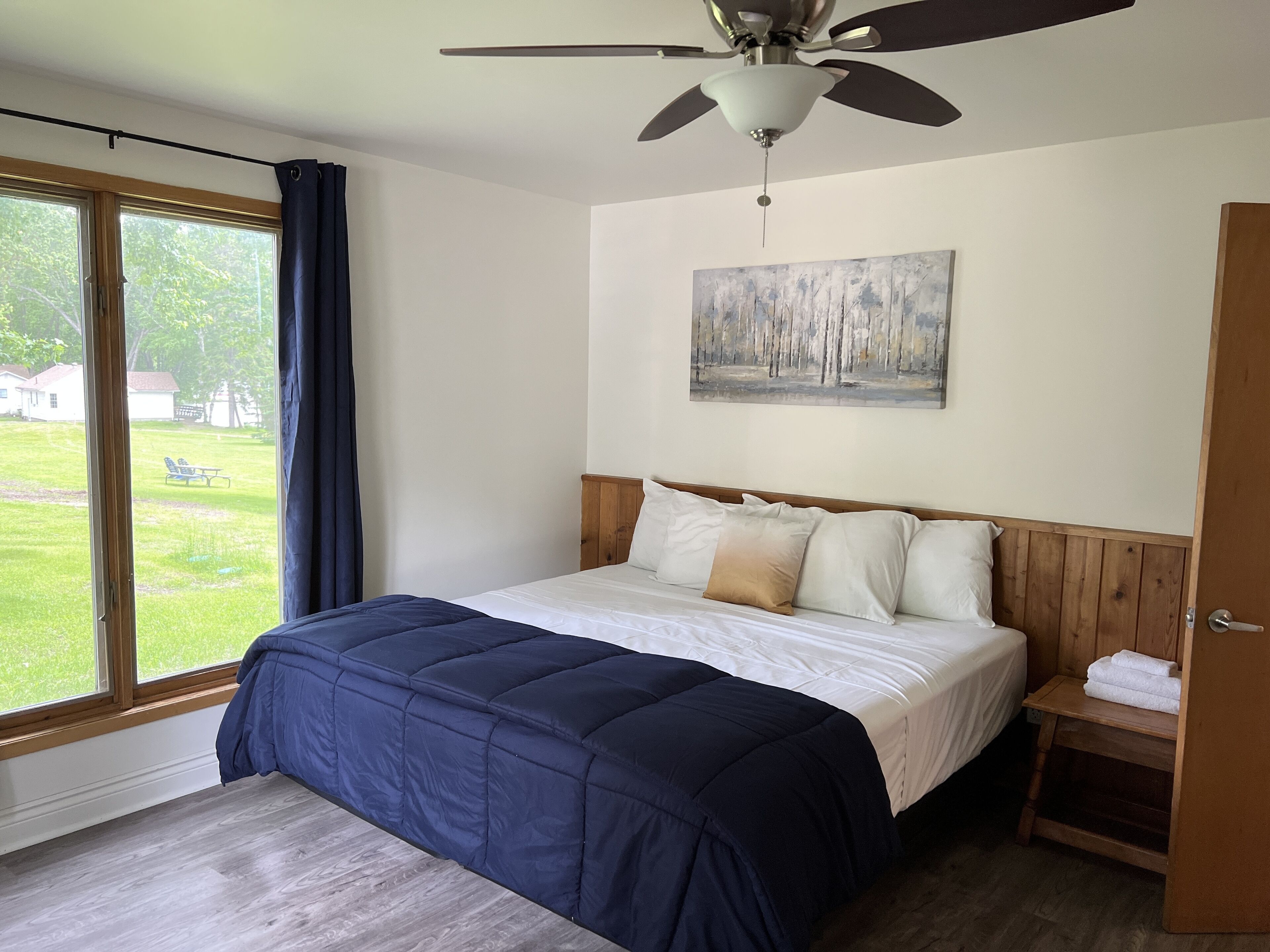 A bedroom with a bed, a ceiling fan, a painting, a window with curtains, and a view of the outside.