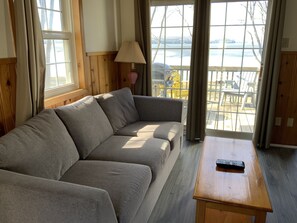 A room with a grey sofa, a wooden table, and a view of a dock and water outside.