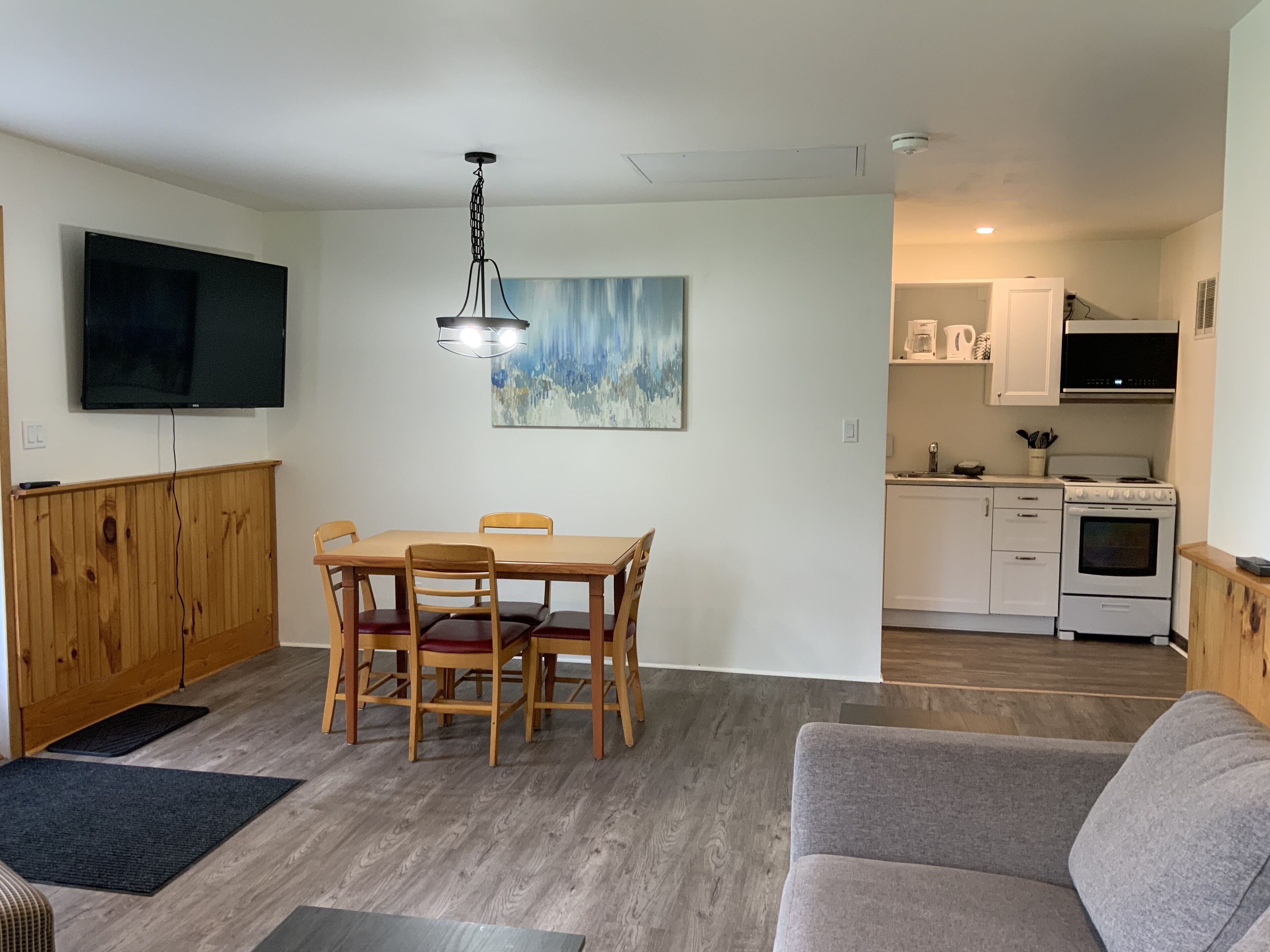 Een moderne keuken met witte kasten, een fornuis en een magnetron. Er is een eethoek met een houten tafel en stoelen. Een flatscreen televisie is aan de muur boven de tafel bevestigd.