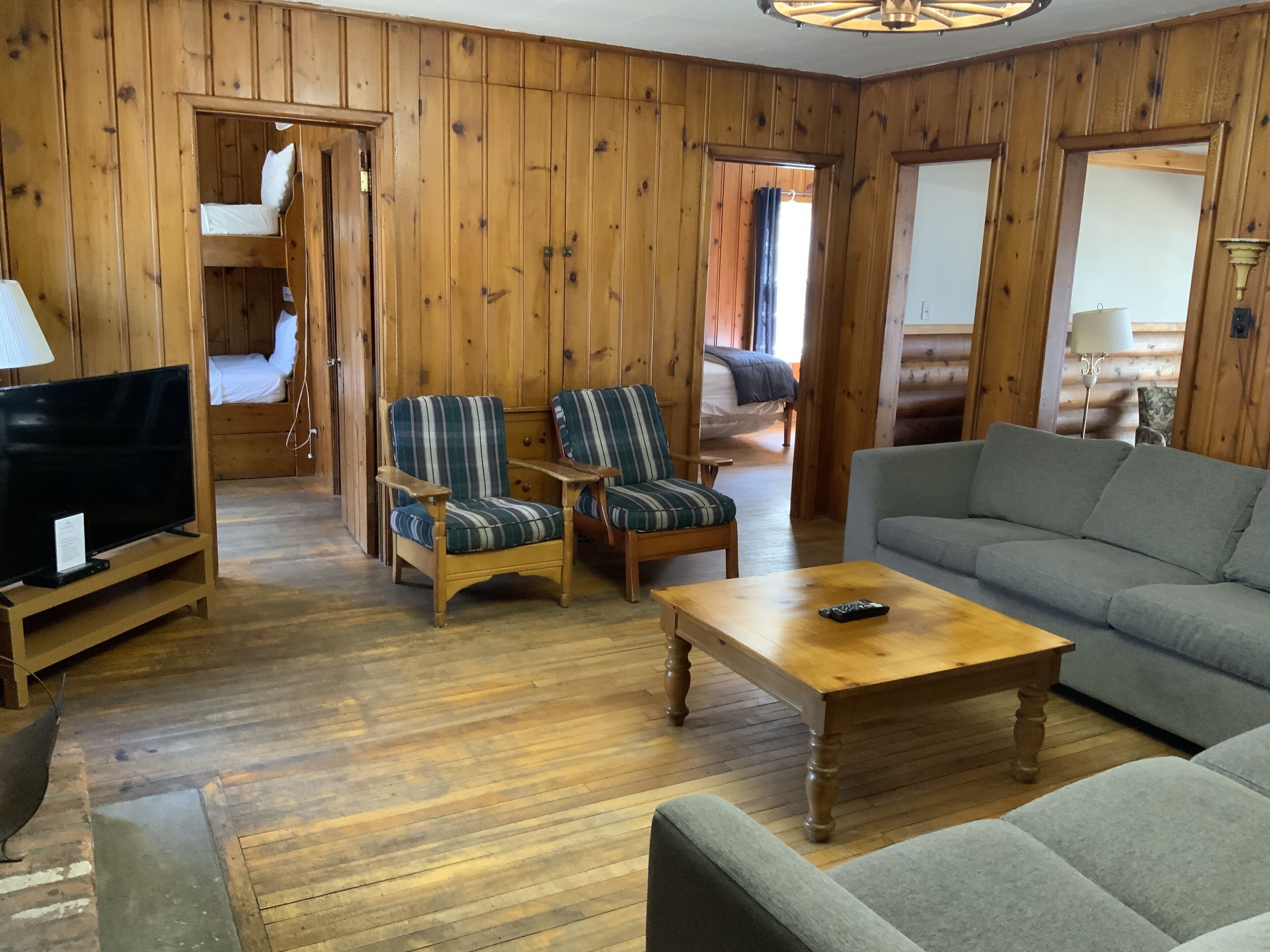 A wooden-paneled room with a television, two striped armchairs, a grey sofa, and a wooden coffee table.