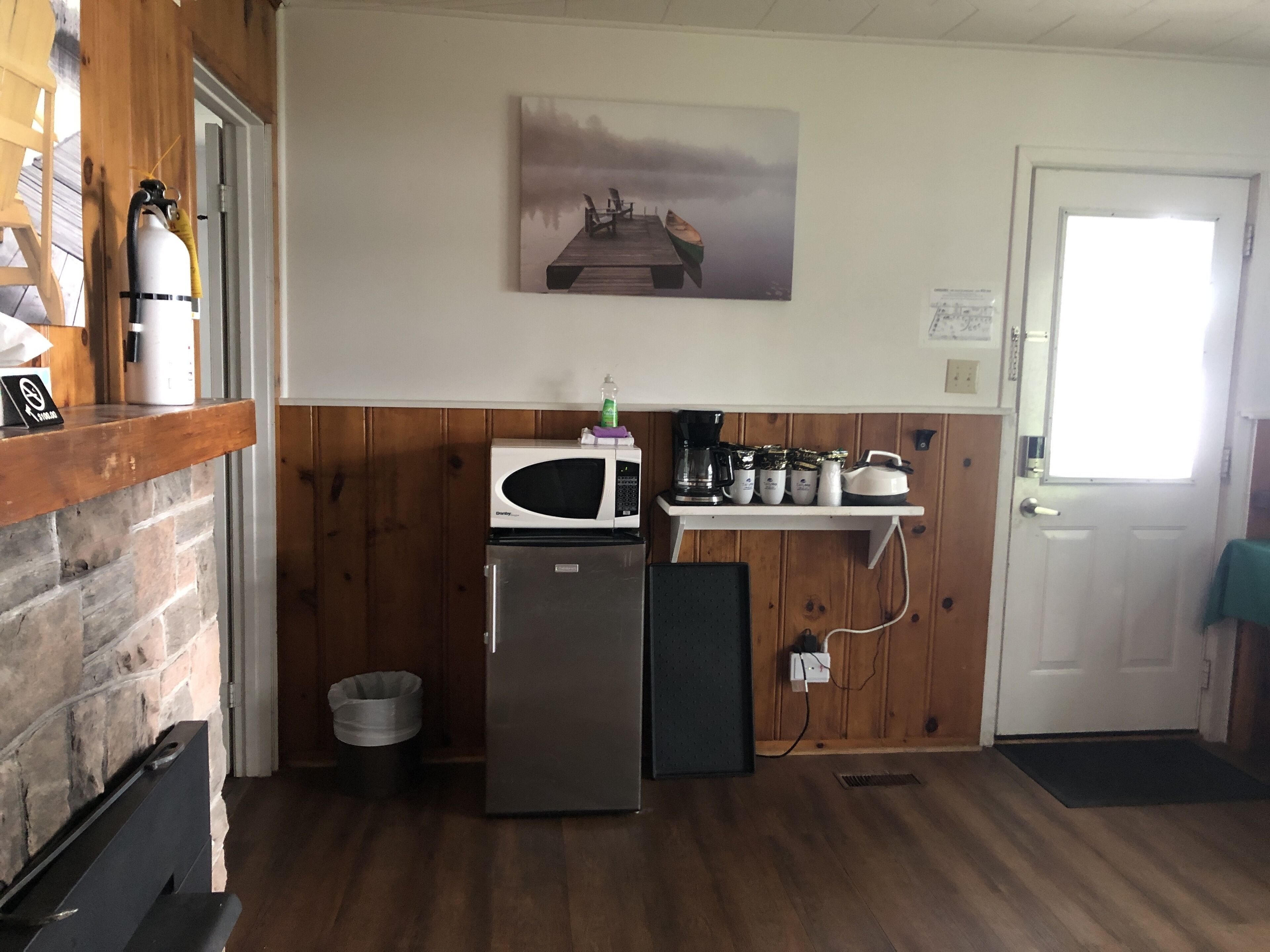 Une cuisine compacte équipée d’un réfrigérateur, d’un four à micro-ondes et d’une machine à café. Une étagère en bois supporte une bouteille d’eau et une petite plante. Un tableau représentant un quai est accroché au mur.