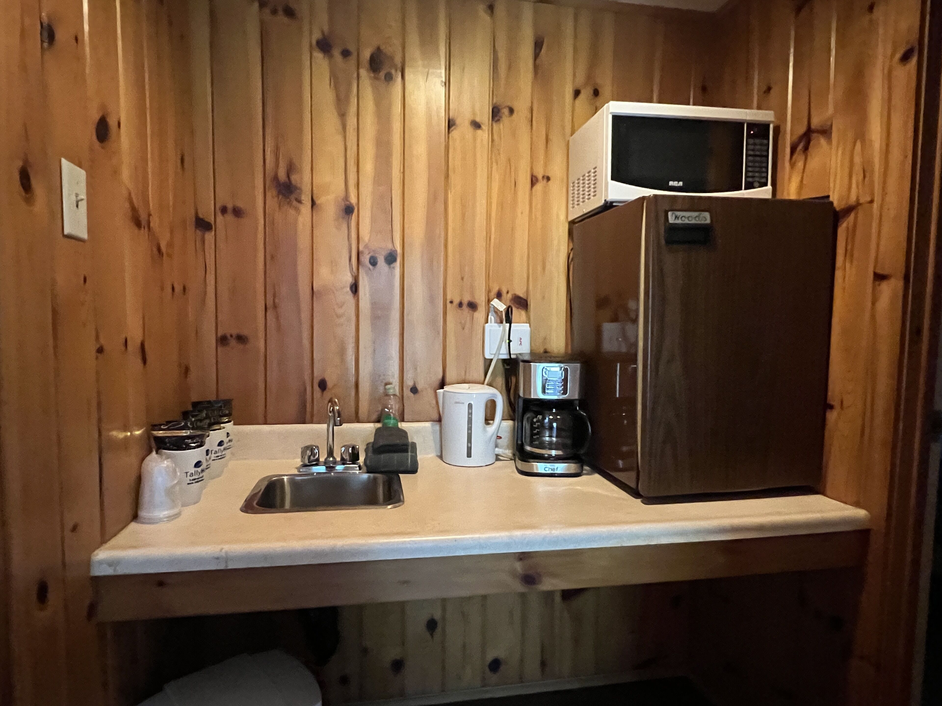 Eine kompakte Küche mit Holzwänden, einem Edelstahl-Spülbecken, einer Kaffeemaschine, einer Mikrowelle und einem Kühlschrank.