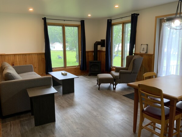 Un salon chaleureux avec un canapé, un pouf, une table et des chaises. On y trouve une cheminée et une vue sur l’extérieur grâce à de grandes fenêtres.