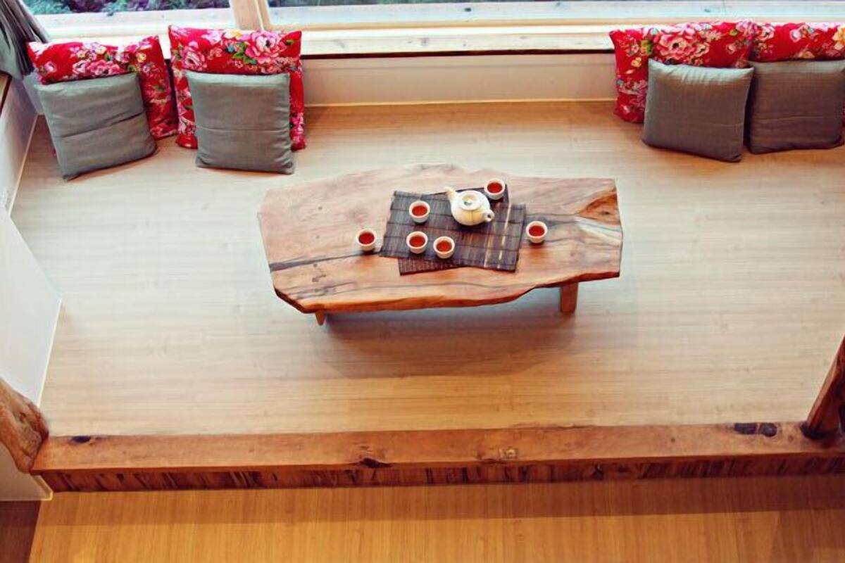 Een houten tafel met een theepot en kopjes, op een houten vloer, omringd door bloemenkussens.