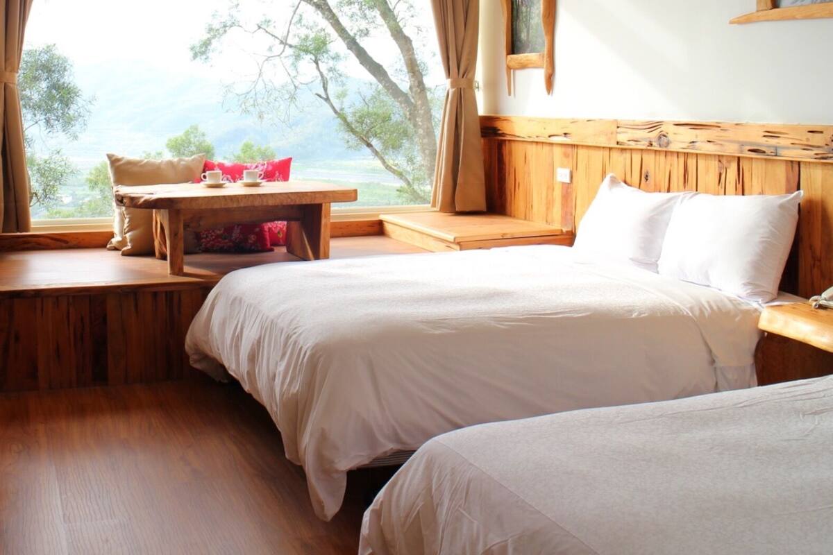 Een slaapkamer met twee bedden, een houten bureau, een tafel met kopje en schotel, en uitzicht op bomen door een raam.