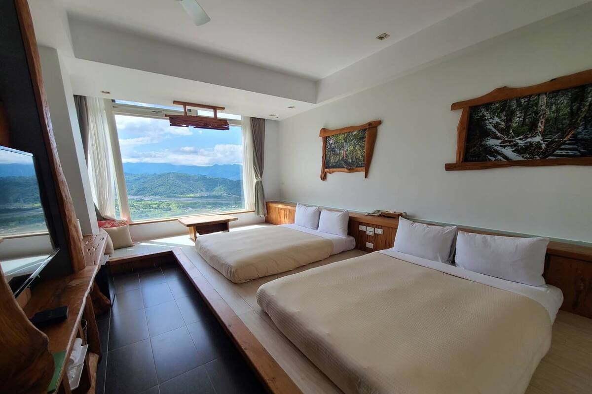 Een hotelkamer met twee bedden, een groot raam met uitzicht op bergen, een schilderij aan de muur en een houten bankje bij het raam.