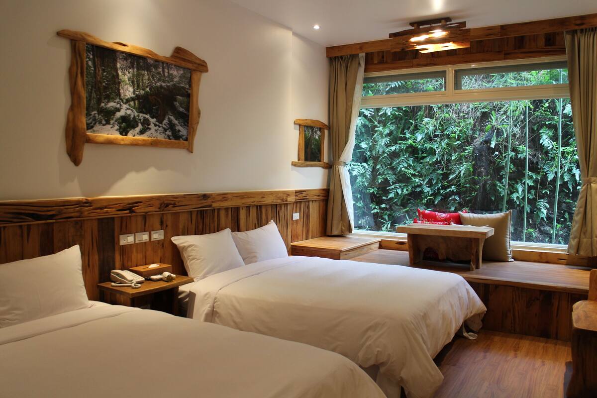 Hotelkamer met twee bedden, een houten hoofdbord, een tafeltje met een rood kussen, een telefoon en een raam met uitzicht op weelderige begroeiing.