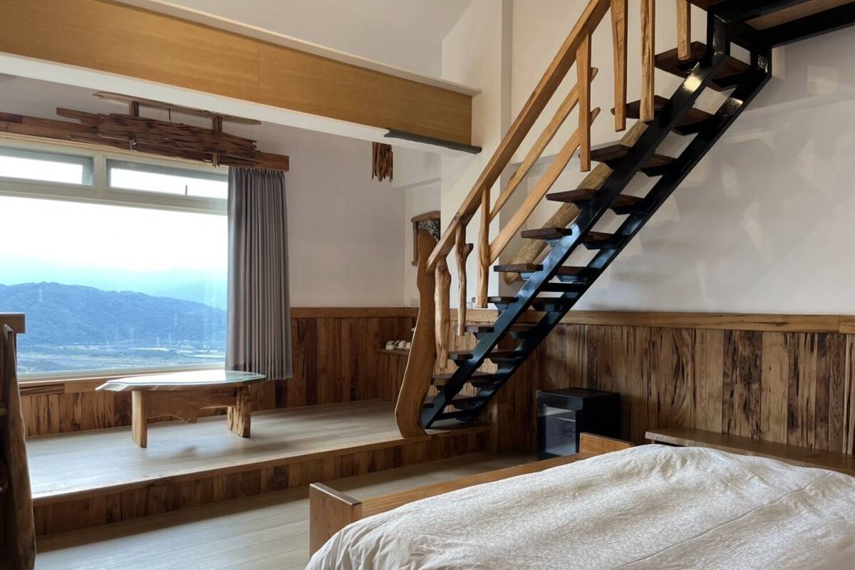 Een slaapkamer met een houten trap, een bed, een raam met uitzicht en een klein tafeltje.