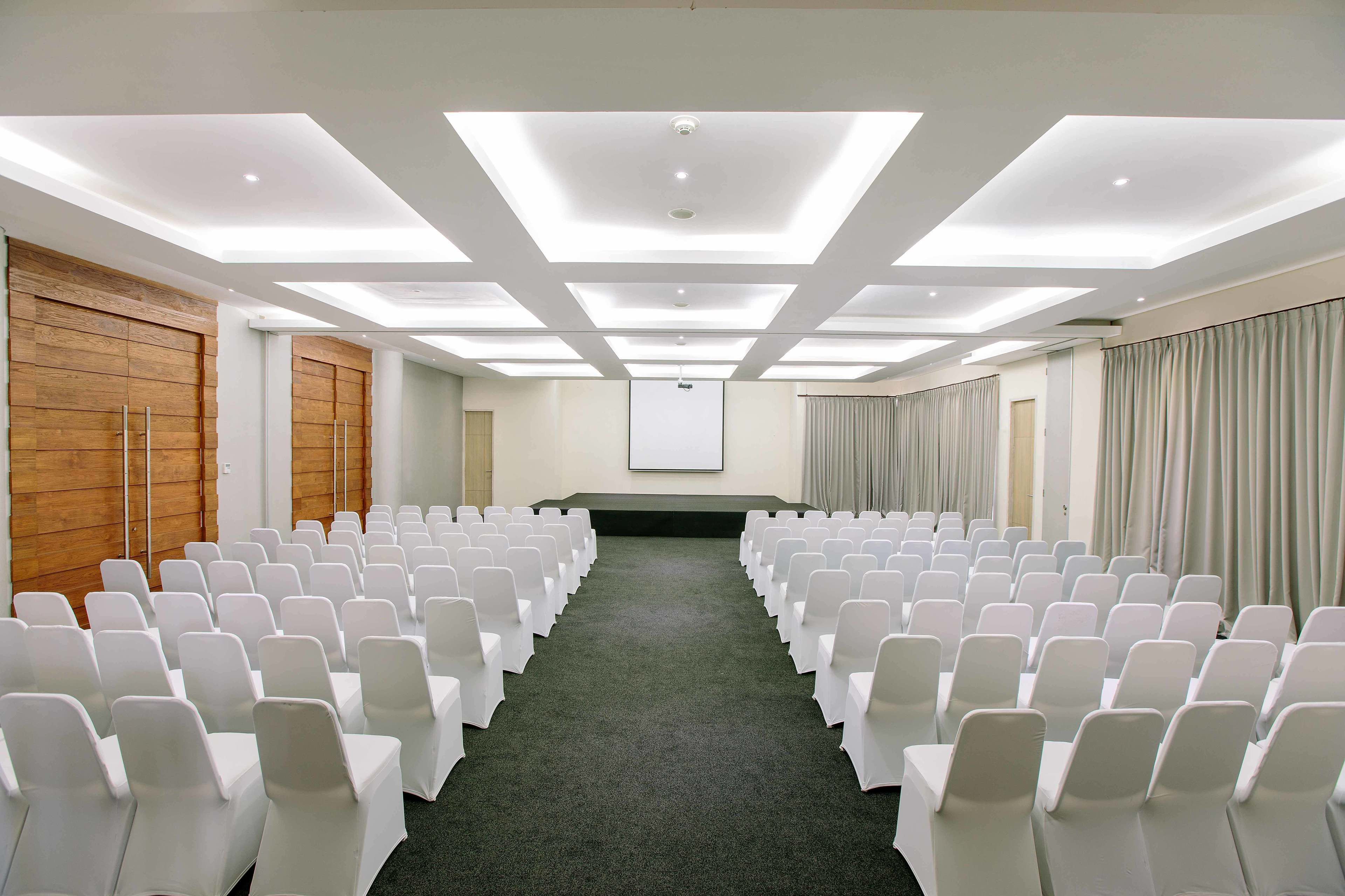 A conference room with rows of white chairs, a projector screen, and a wooden panel wall.