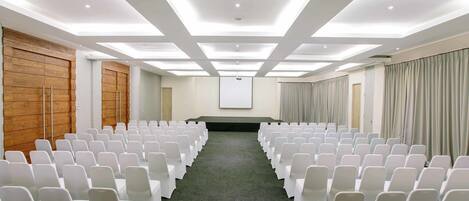 A conference room with rows of white chairs, a projector screen, and a wooden panel wall.