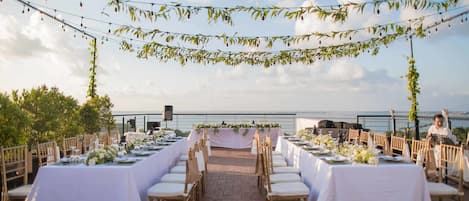 Montaje para una boda al aire libre con mesas y sillas dispuestas para los invitados, un micrófono y vista al mar de fondo.