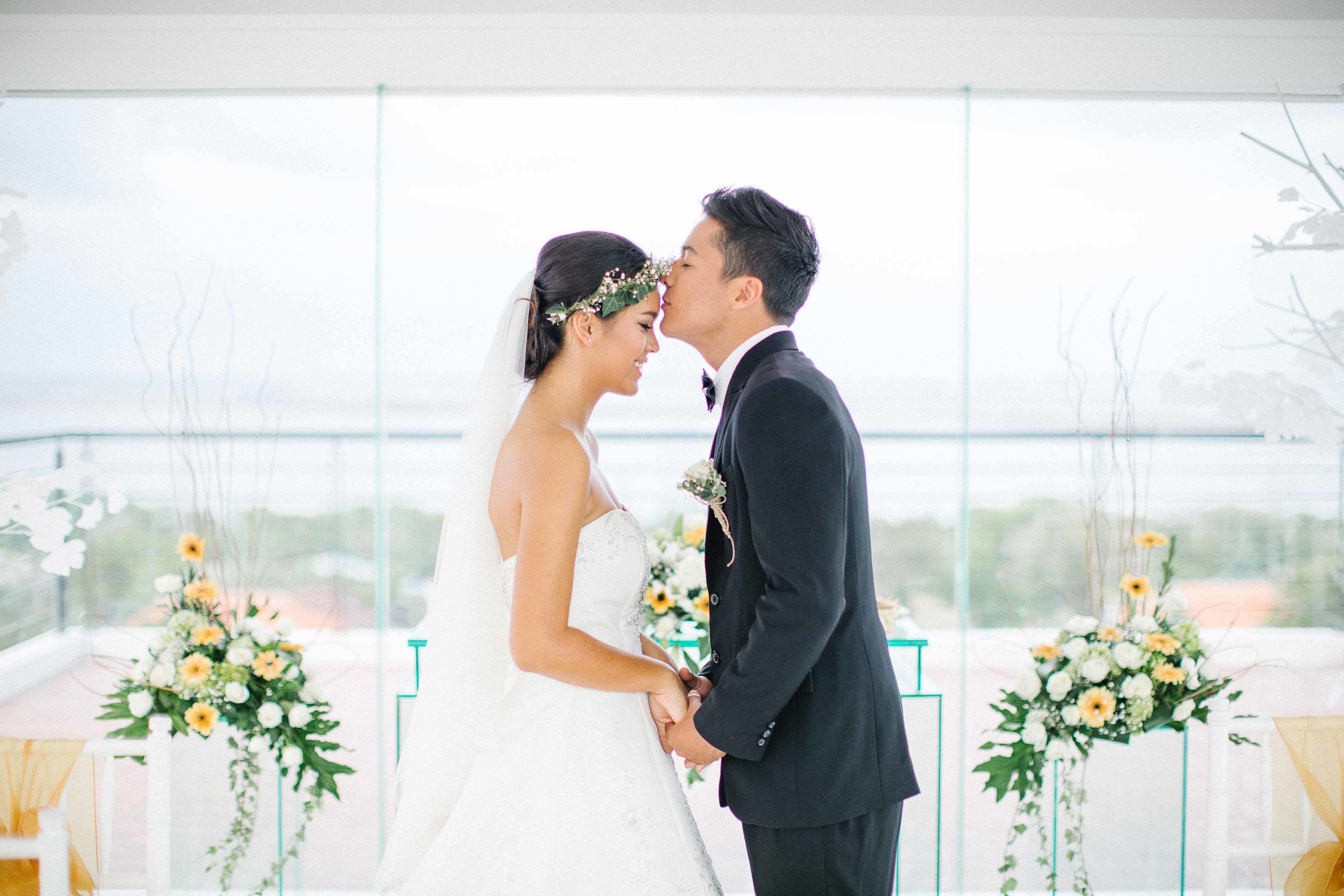 A couple in wedding attire standing close together, with floral arrangements and a balcony view in the background.
