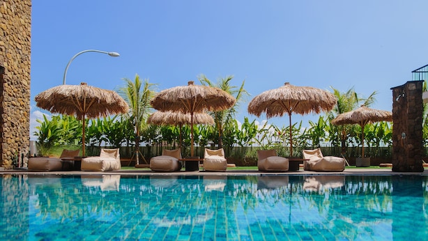 A swimming pool with clear blue water, surrounded by lounge chairs and thatched umbrellas, with a stone wall and palm trees in the background. - Mahogany Hotel (Nusa Dua)