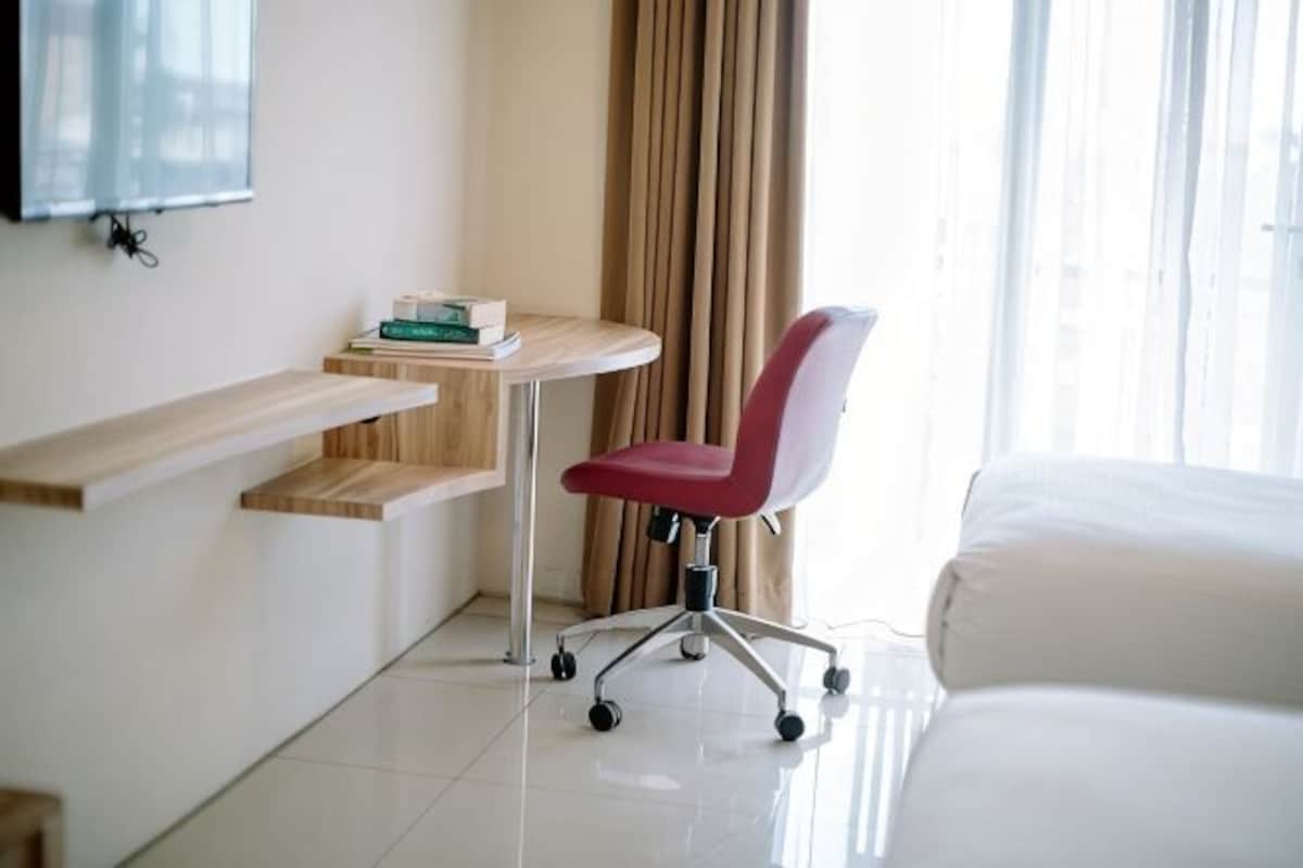 A hotel room with a bed, a desk with a chair, a TV mounted on the wall, and a window with curtains.