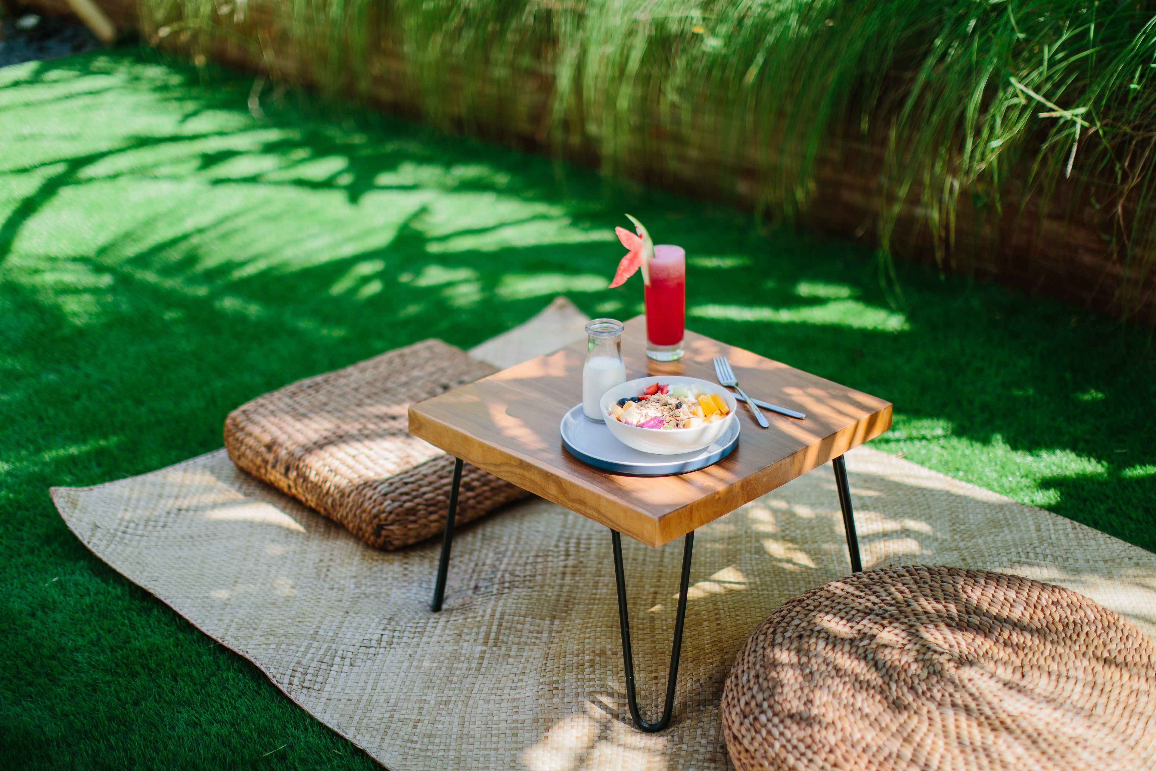 A picnic setup with a table, a bowl of food, a drink, and a bottle of water on a mat in a grassy area.