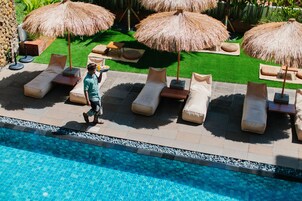 Zona junto a la piscina con sillones de descanso y sombrillas de paja, césped cuidado y piscina de aguas cristalinas.
