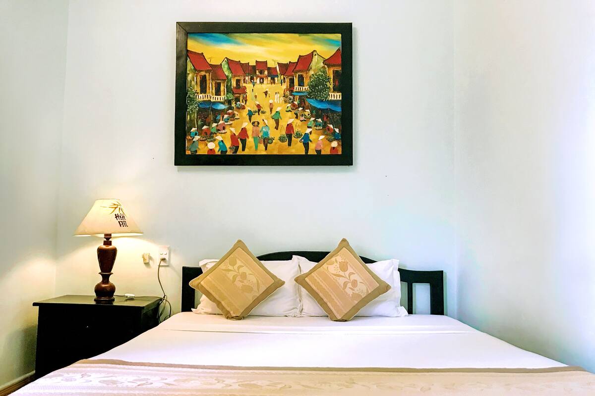 A hotel room with a bed, two decorative pillows, a nightstand with a lamp, and a painting of a village scene on the wall.