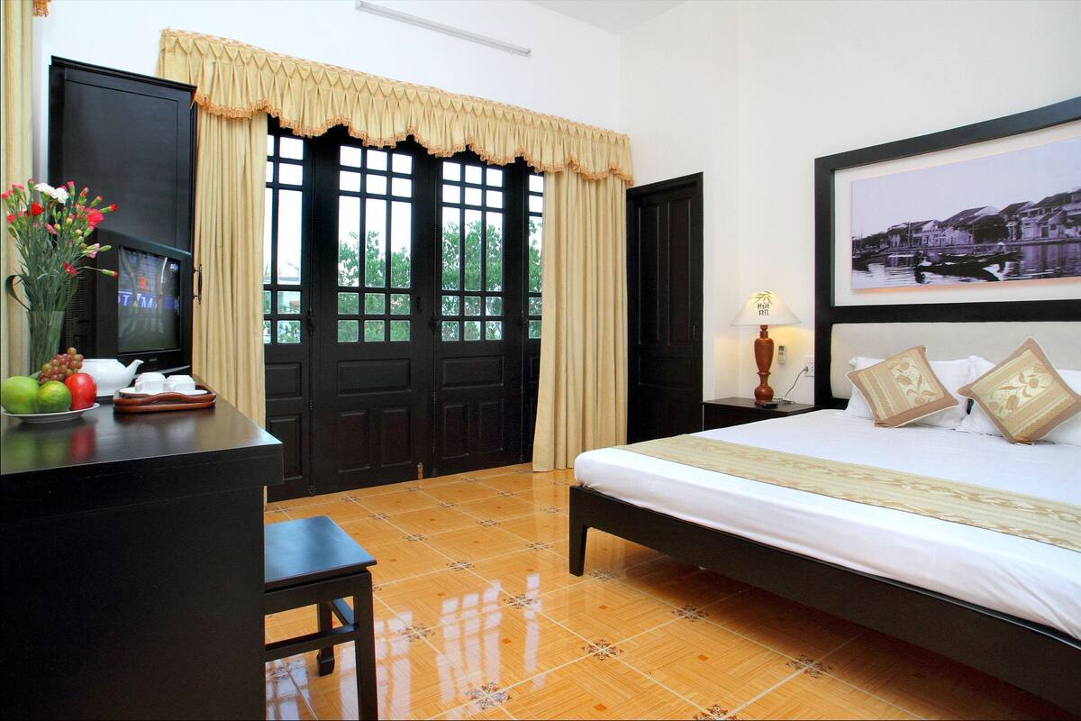 A hotel room with a bed, a desk with a television, a vase with flowers, and a framed picture on the wall.