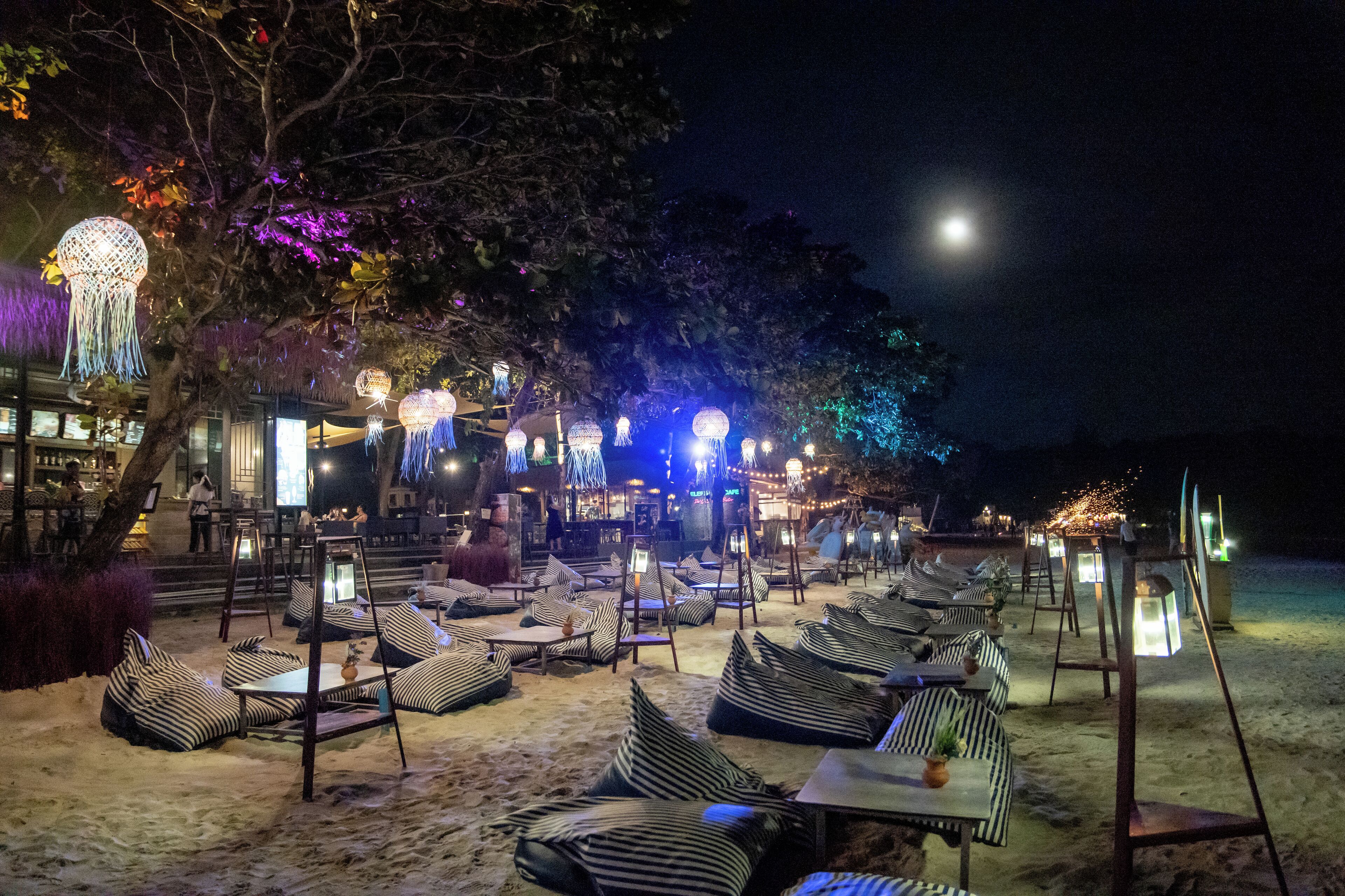 Un espace extérieur aménagé avec des sièges, des coussins à rayures et des tables, entouré d’arbres et éclairé par des guirlandes lumineuses suspendues.