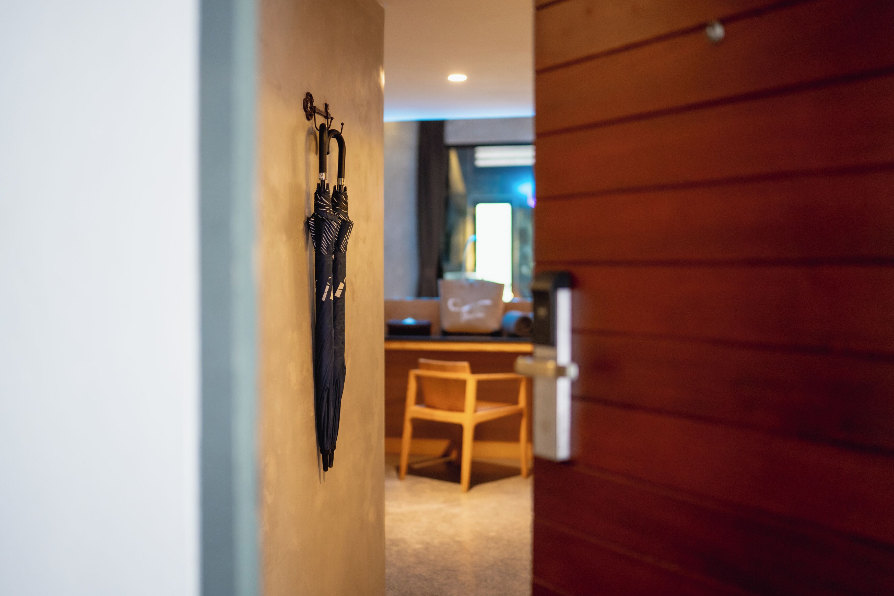 A hallway with a wooden door, a wall-mounted umbrella, a wooden chair, and a desk with a bag and a computer.