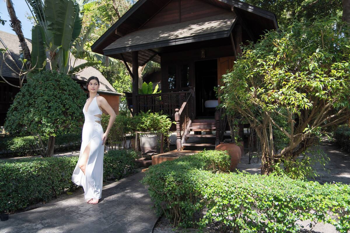 A woman in a white dress standing in a garden with a wooden house in the background.