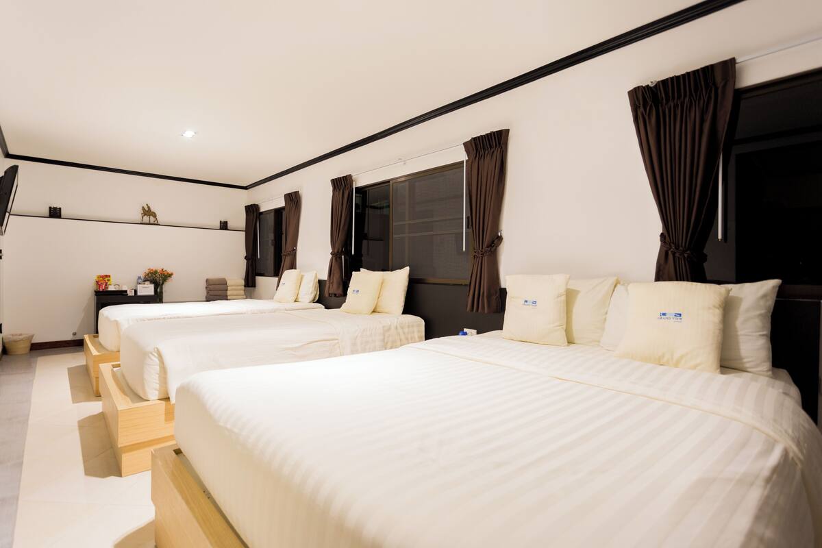 A hotel room with three beds, a flat-screen TV, a small table with a vase, and a window with curtains.