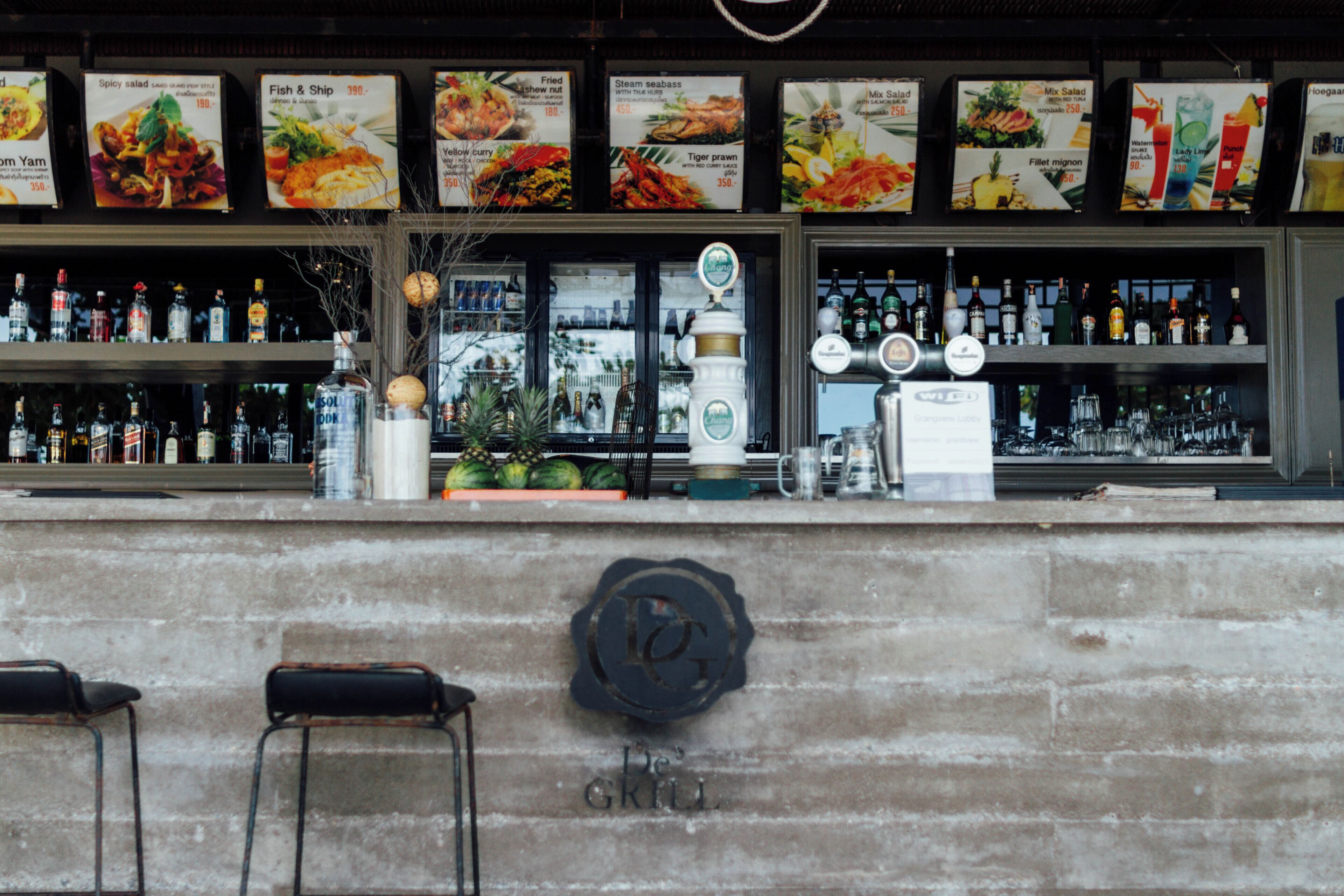 A bar area with a menu board displaying various dishes, a selection of bottles, and a counter with stools.