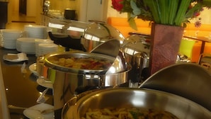 Un área de buffet con múltiples estaciones de servicio, incluyendo pasta y otros platos. Hay pilas de platos y cuencos, y una planta en maceta con flores.