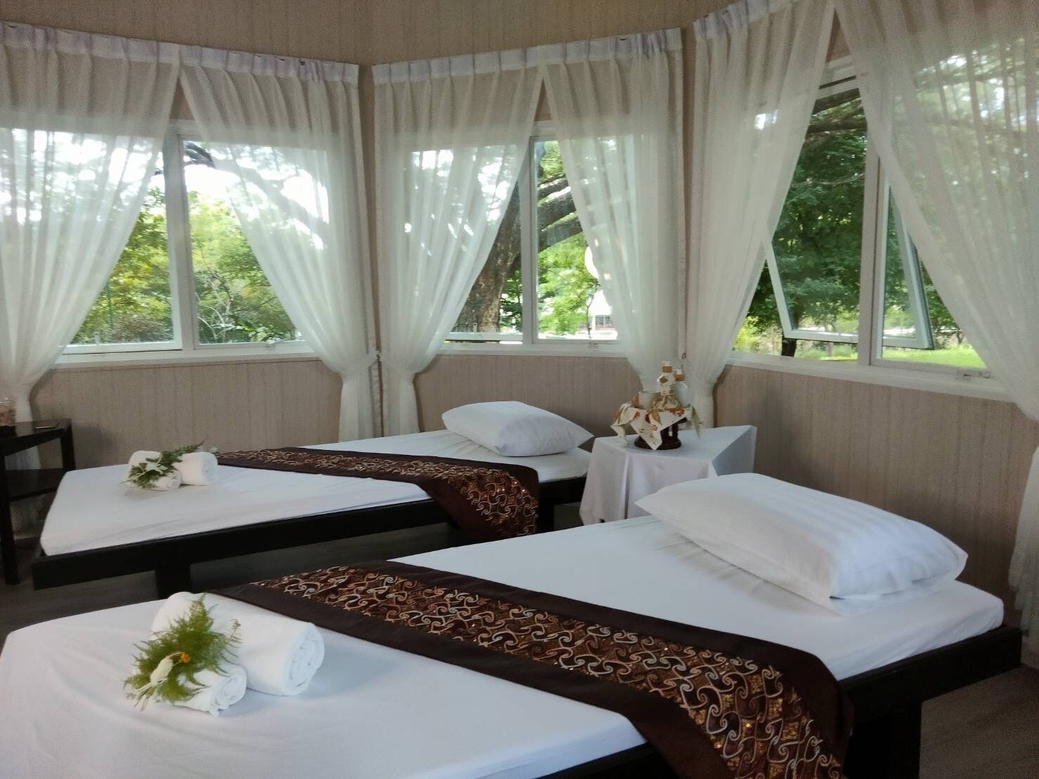 Habitación con dos camas, cada una con cubrecama blanco y cabecero marrón decorativo. Hay cortinas blancas en las ventanas, y una pequeña mesa con un jarrón de flores entre las camas.