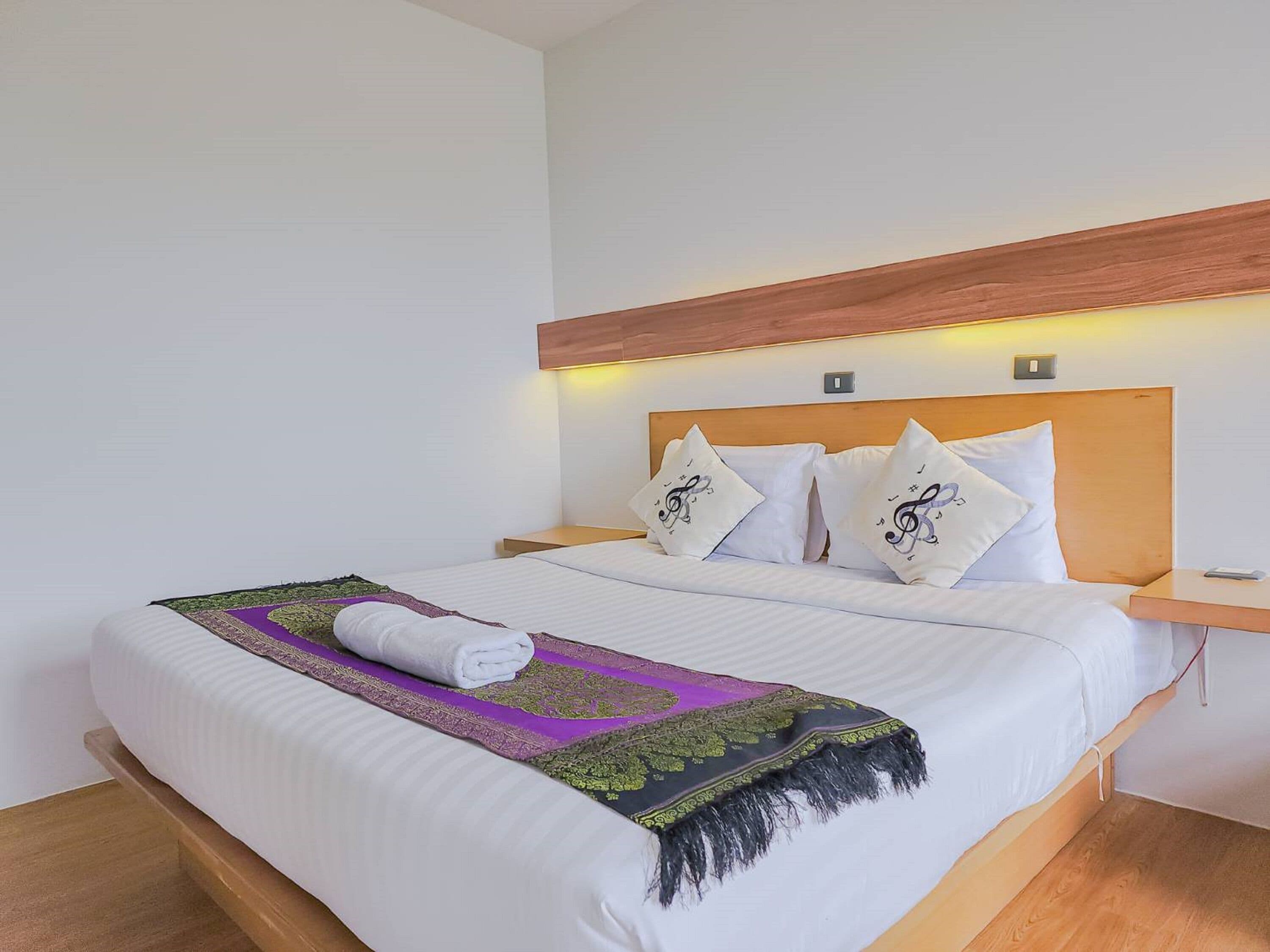 A neatly made bed with a white comforter, a rolled white towel, and a decorative throw blanket. There are two bedside tables with lamps, and the room has a wooden headboard and a white wall.
