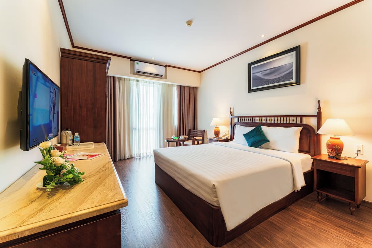 Ein Hotelzimmer mit Bett, Nachttisch, Lampe, Fernseher und Schreibtisch mit Blumenvase.